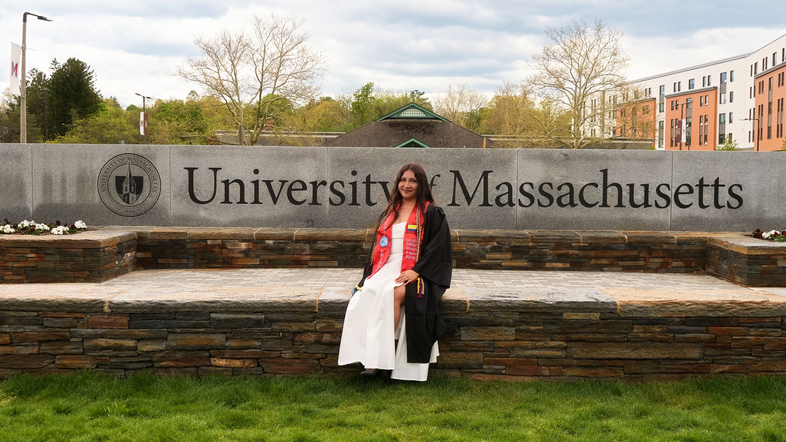 Graduate in cap and gown poses proudly in front of University of Massachusetts sign on a cloudy spring day.