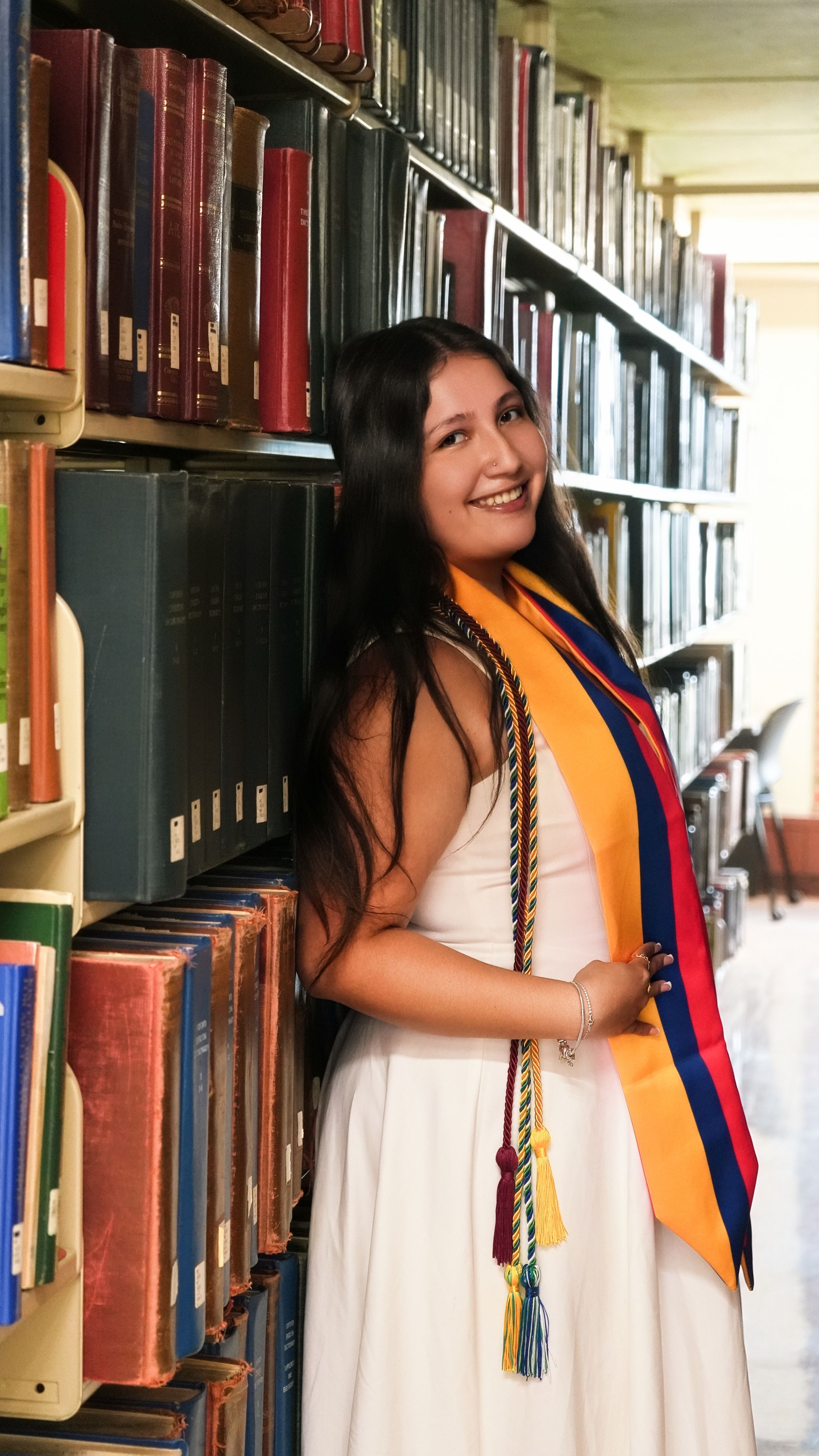 Smiling graduate in white dress with orange and blue sash poses by library bookshelves.