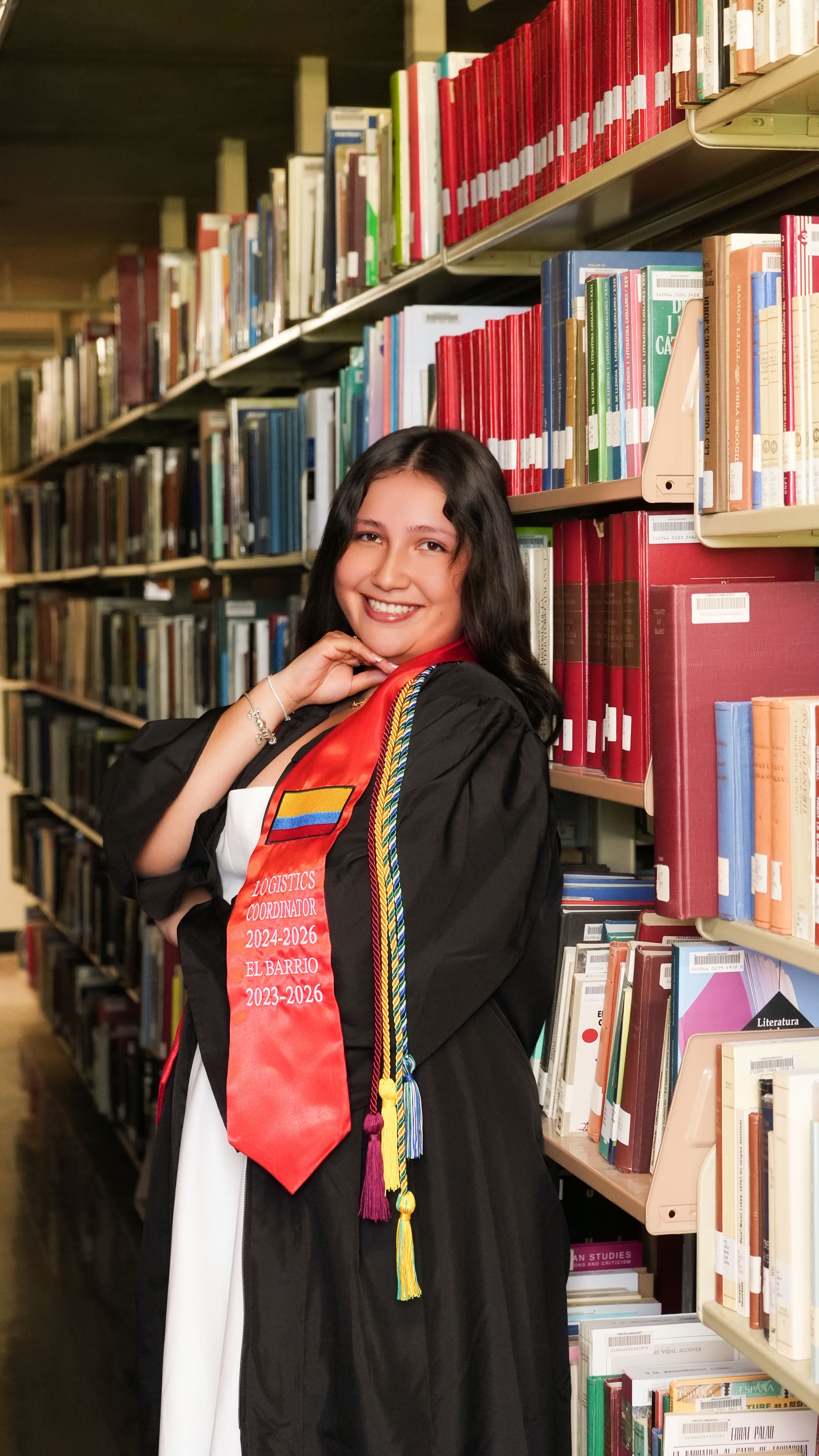 Graduate in black gown and red stole smiles in library aisle surrounded by colorful books.
