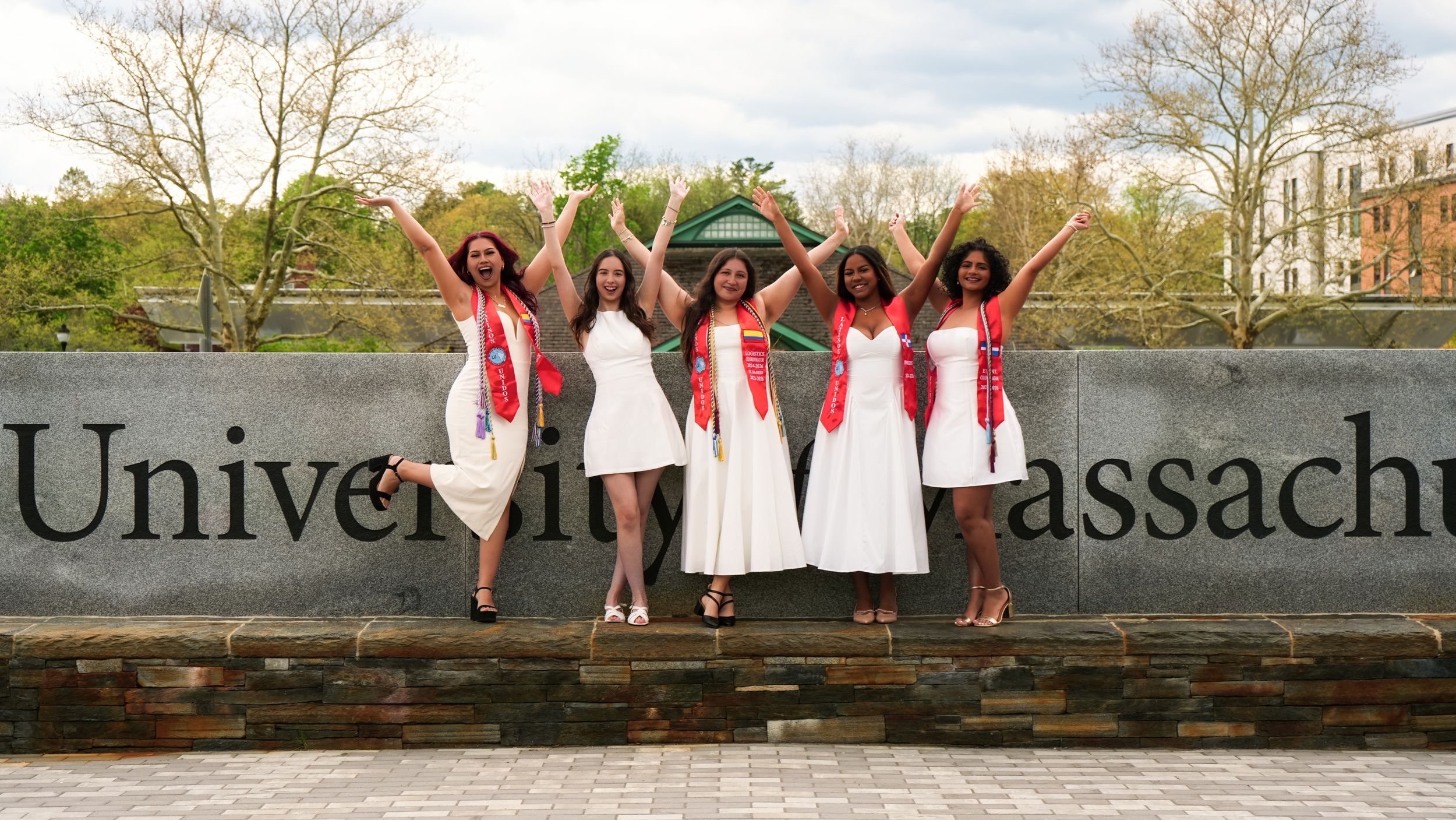 Five graduates in white dresses and red sashes jump joyfully in front of University of Massachusetts sign outdoors.