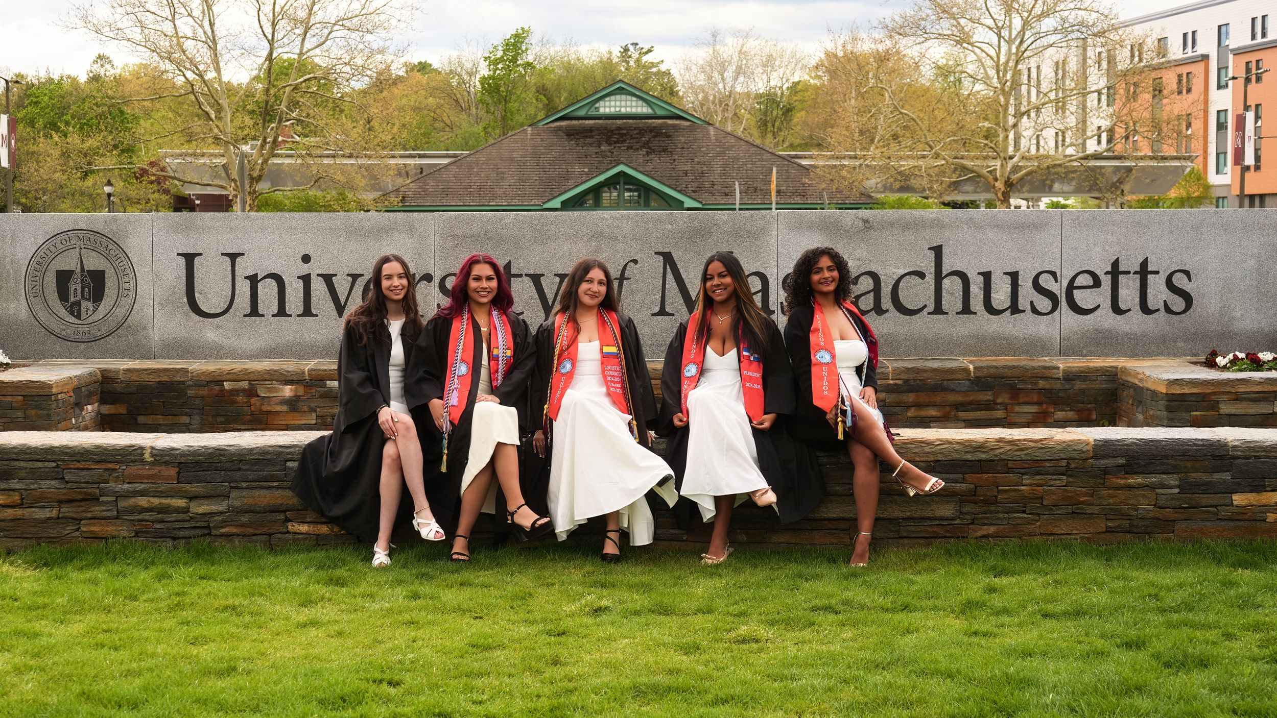 Five graduates in caps and gowns pose in front of University of Massachusetts sign on a cloudy spring day.