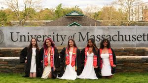 Five graduates in black robes with red sashes pose proudly in front of the University of Massachusetts sign.