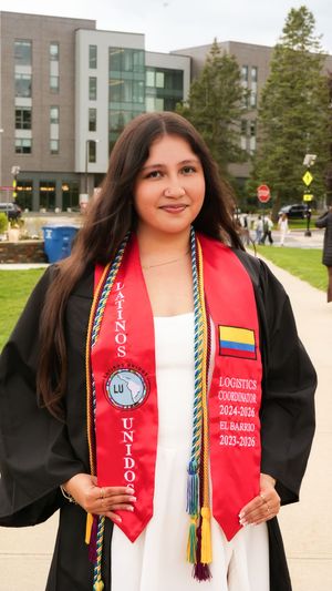 A Latina college graduate smiles in black graduation gown with Latinos Unidos and Colombian flag stoles on campus.