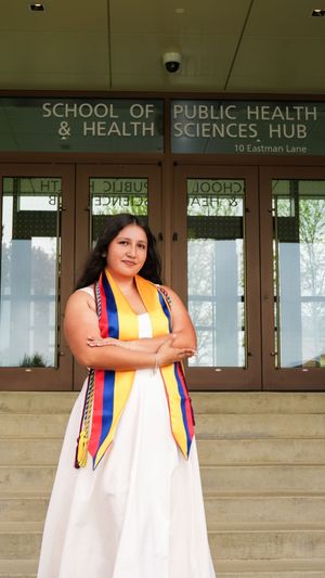 Graduate in white gown with colorful stole stands proudly before School of Public Health & Health Sciences Hub building.