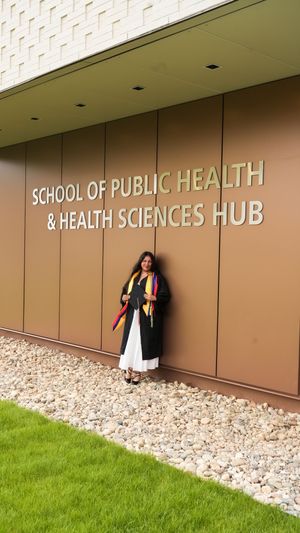Graduate in black gown stands before School of Public Health & Health Sciences Hub building.
