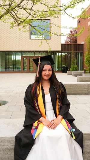 A young woman in black graduation cap and gown with colorful stoles sits outdoors on a stone bench near a modern building.