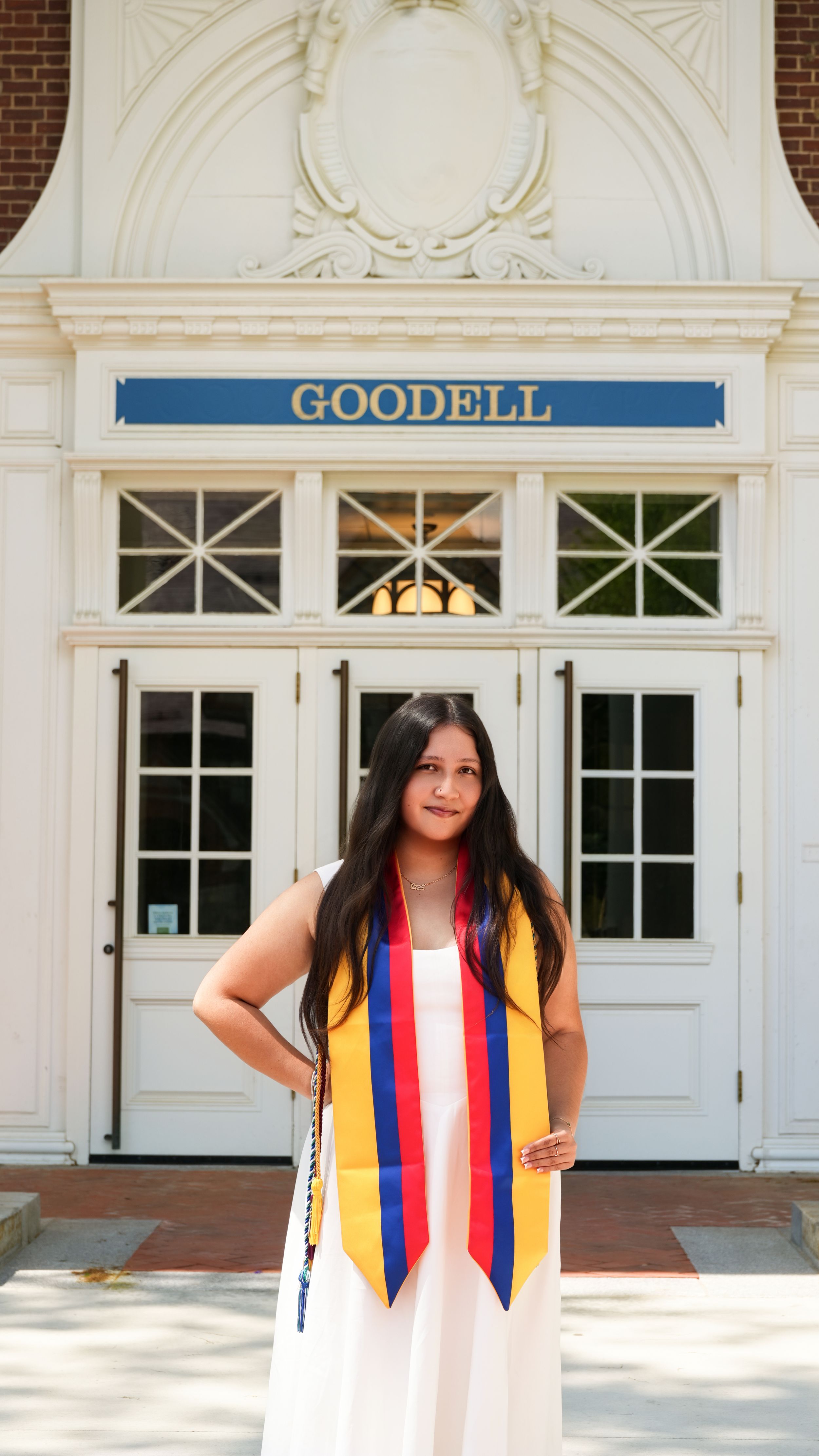 A graduate poses in a white gown with colorful stole in front of Goodell building on a sunny day.