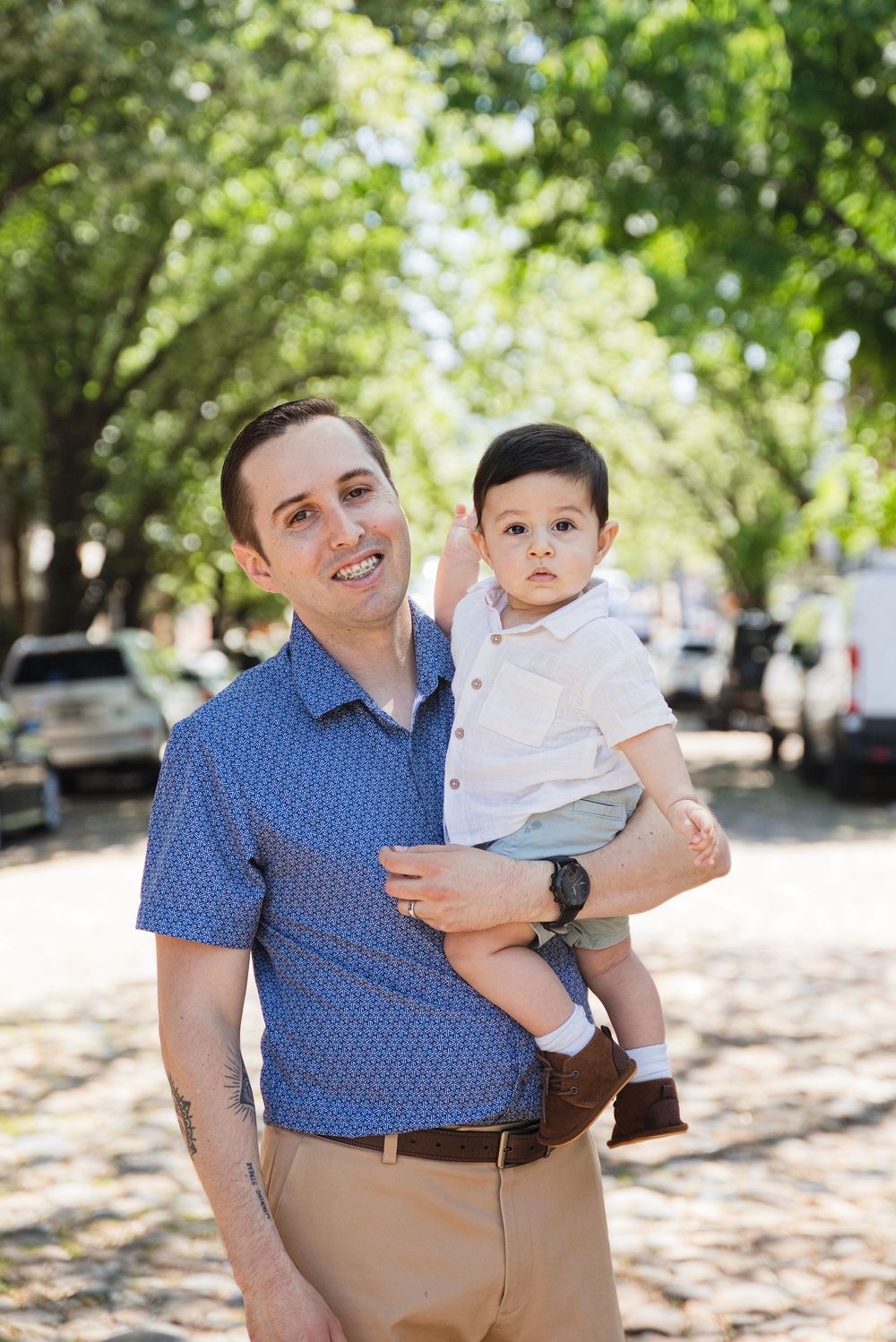 A smiling man in a blue shirt holds a young toddler boy on a tree-lined cobblestone street.
