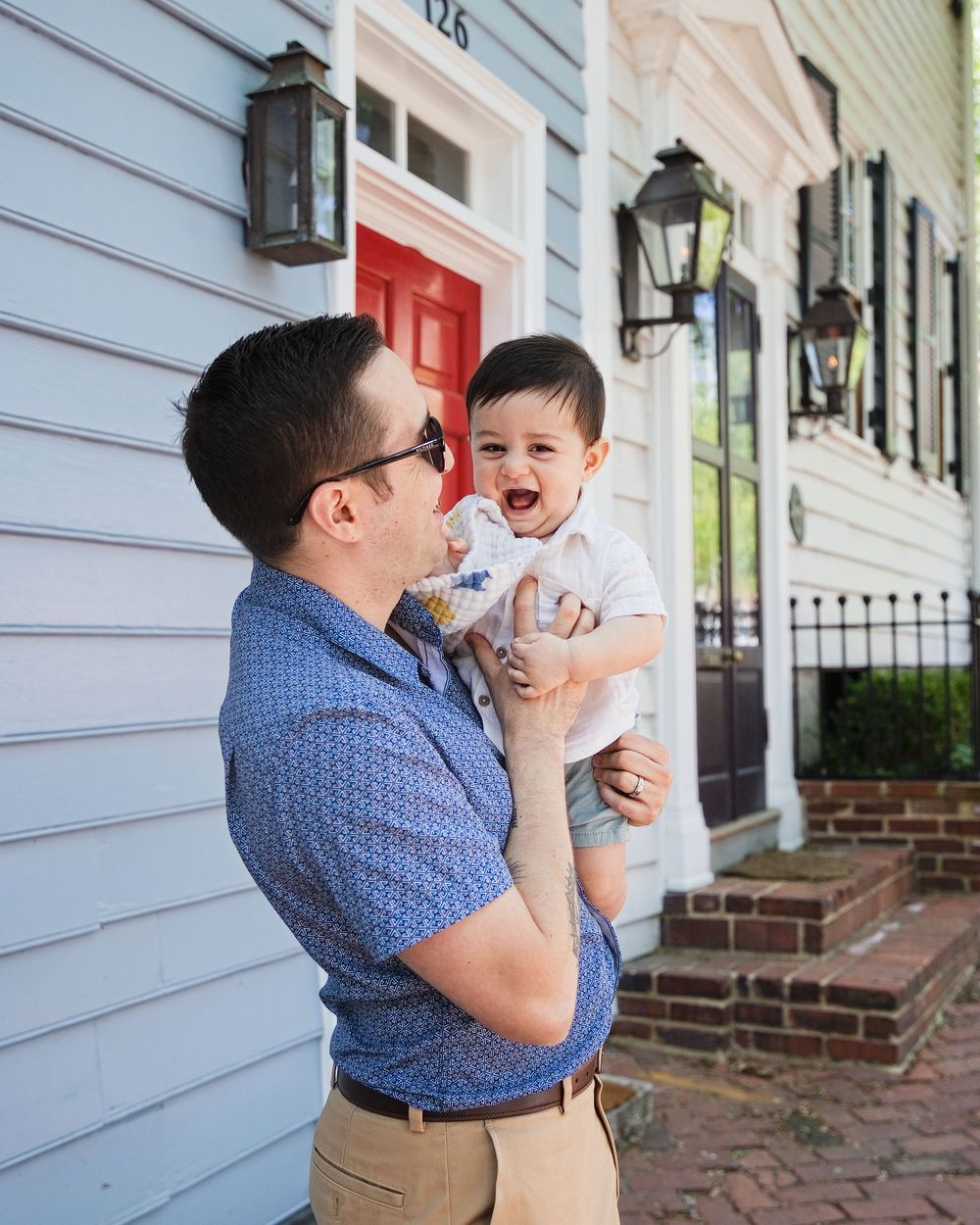A man in a blue polo shirt holds a laughing toddler outside a charming home with a red door.