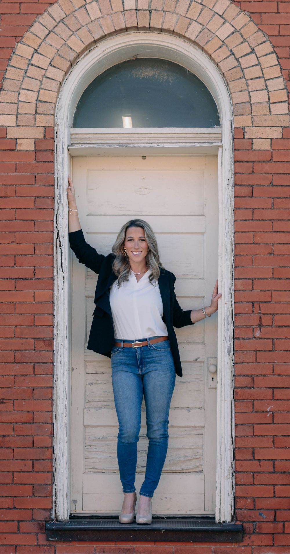 Woman in blazer and jeans posing confidently in an arched white doorway of a red brick building.