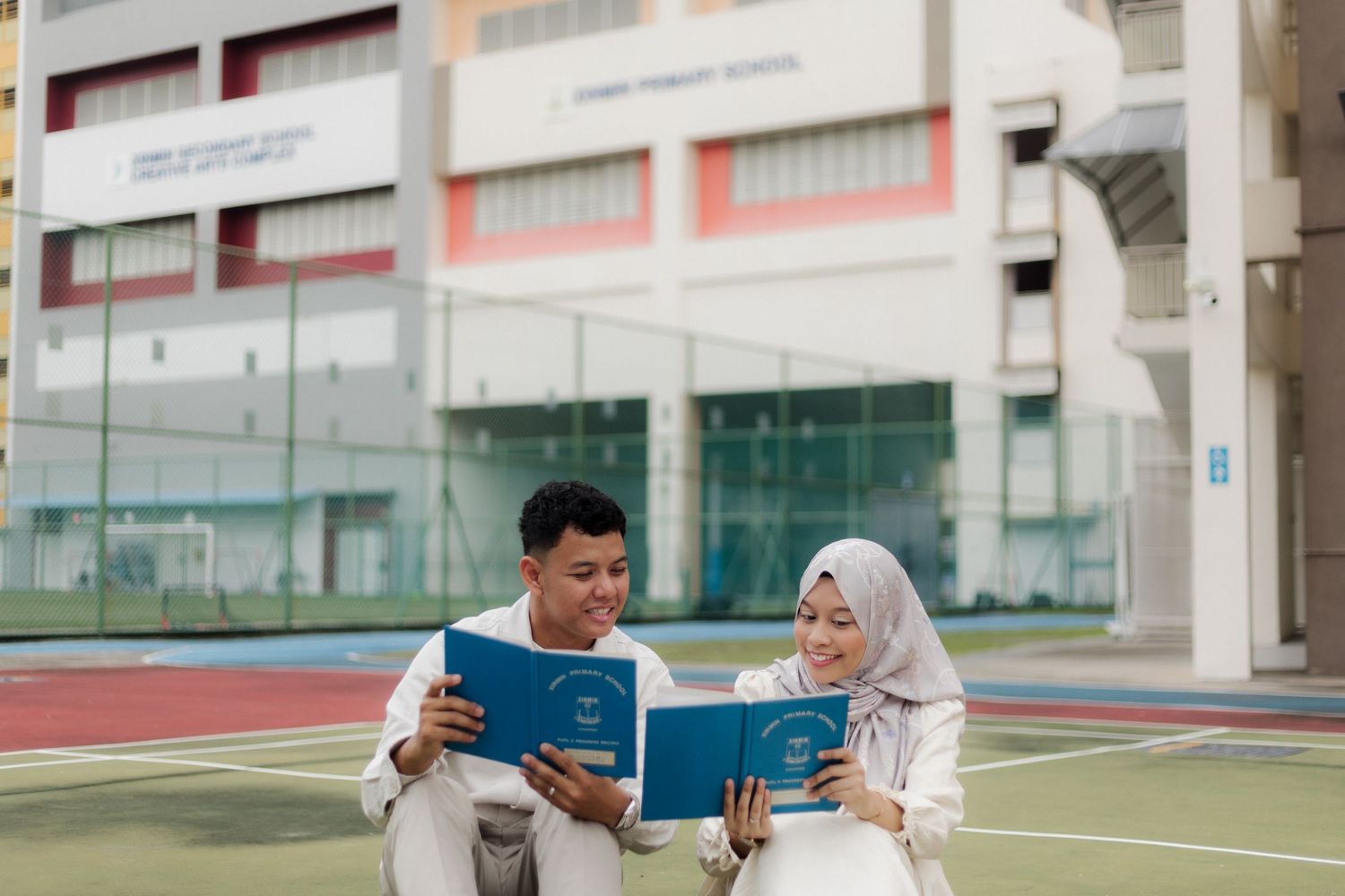 Muslim couple in white attire reading blue books on a school sports court outdoors.