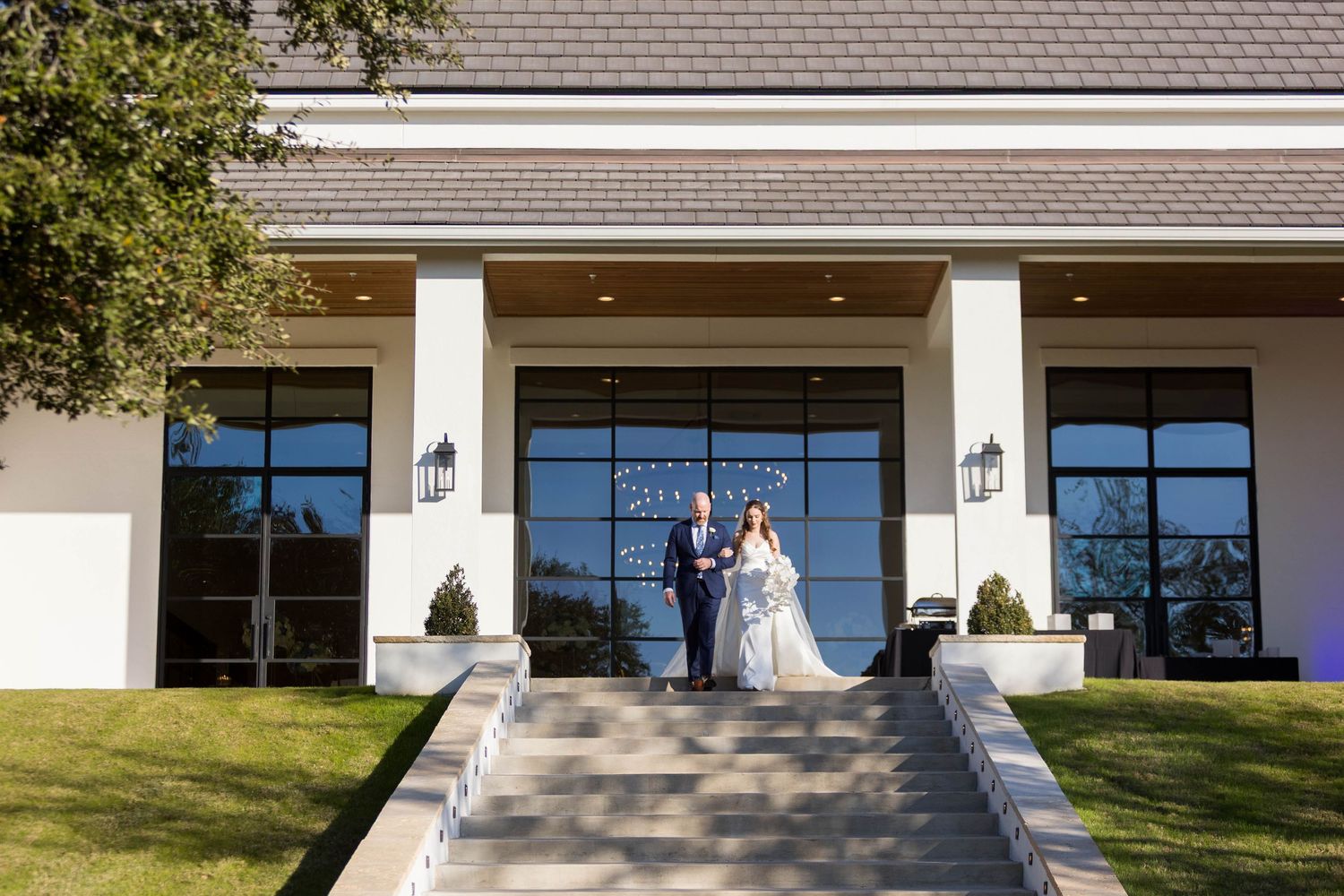 Bride and groom pose on grand entrance steps of elegant white wedding venue with large windows.