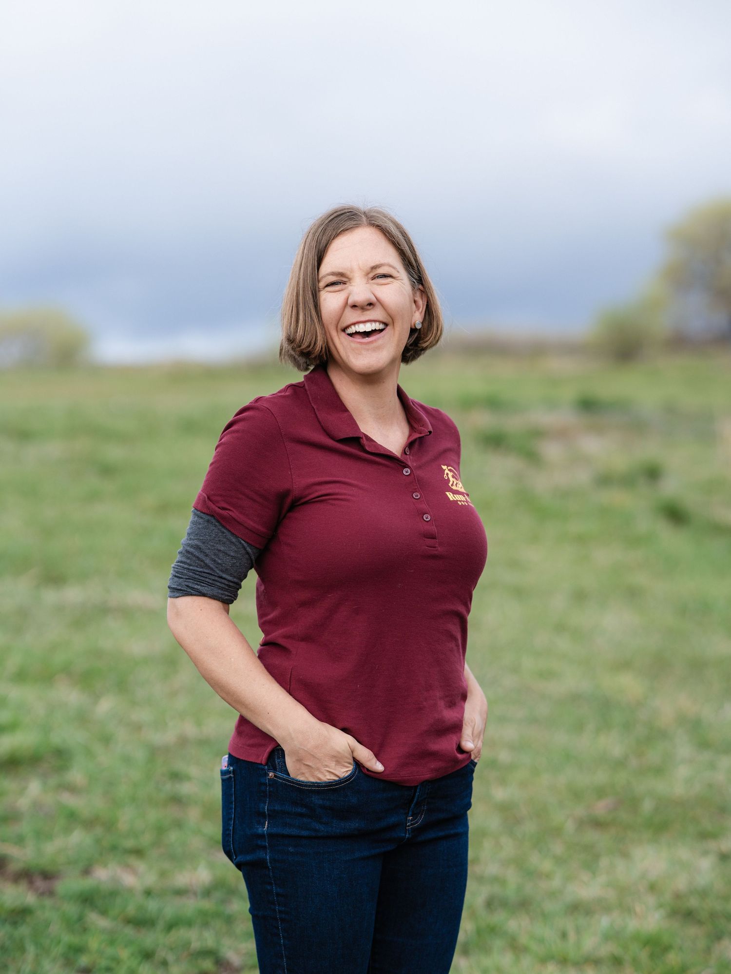 Woman in maroon polo kneeling beside a Rhodesian Ridgeback dog in a purple wildflower field with hills in background.