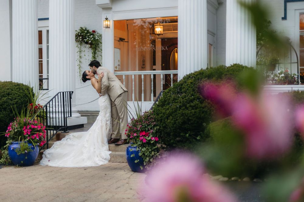 Bride and groom share a romantic kiss on the steps of a white columned venue Meson SAbika in Naperville surrounded by colorful flowers.