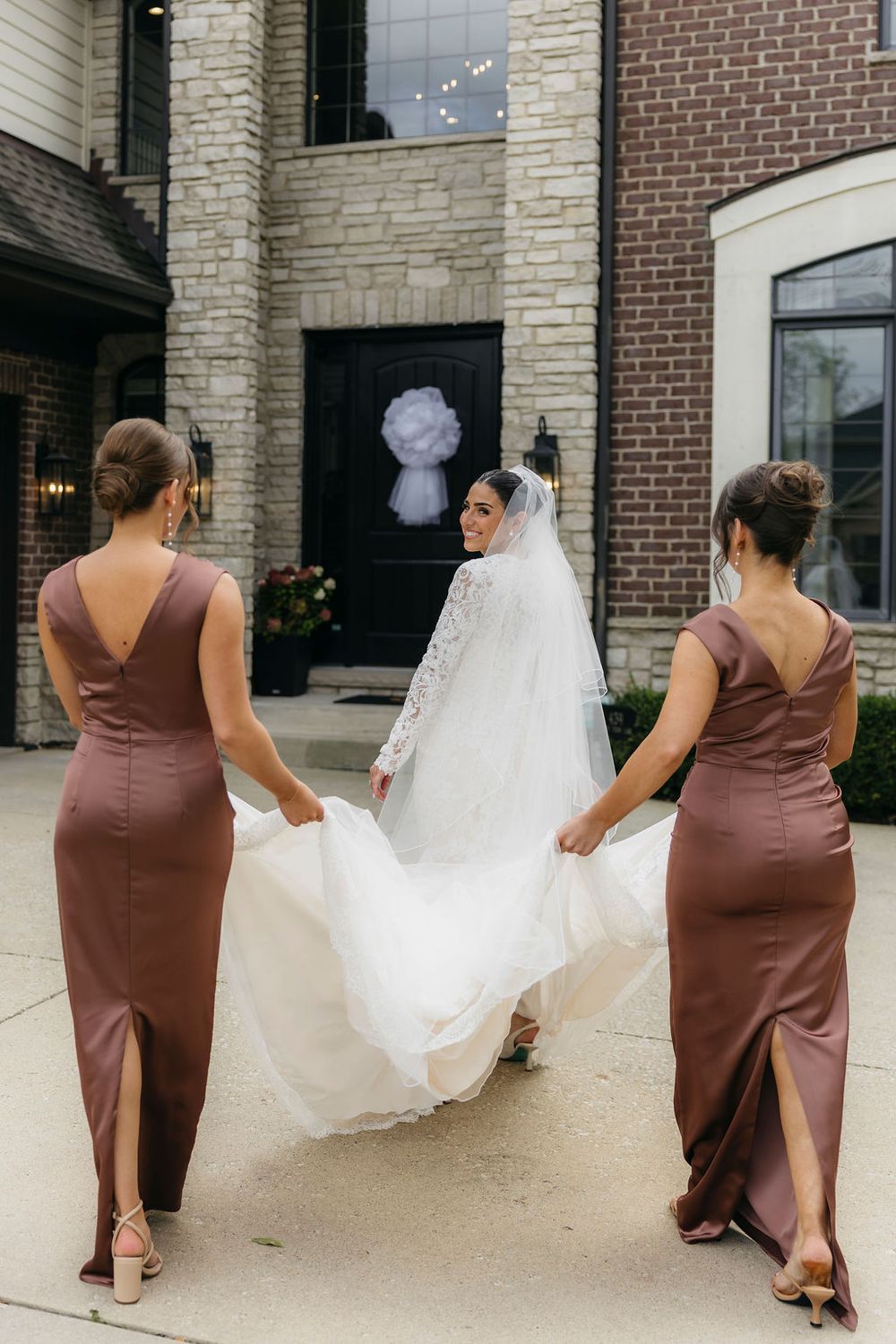 Bride in white lace gown and veil walks with two bridesmaids in mauve satin dresses holding her train in Chicago