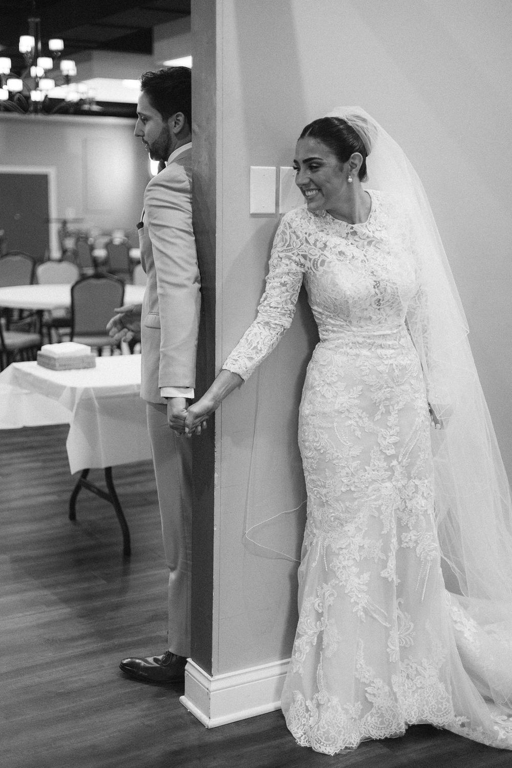 Black and white photo of bride in lace gown and veil holding hands with groom around a pillar at church before the ceremony in Chicago