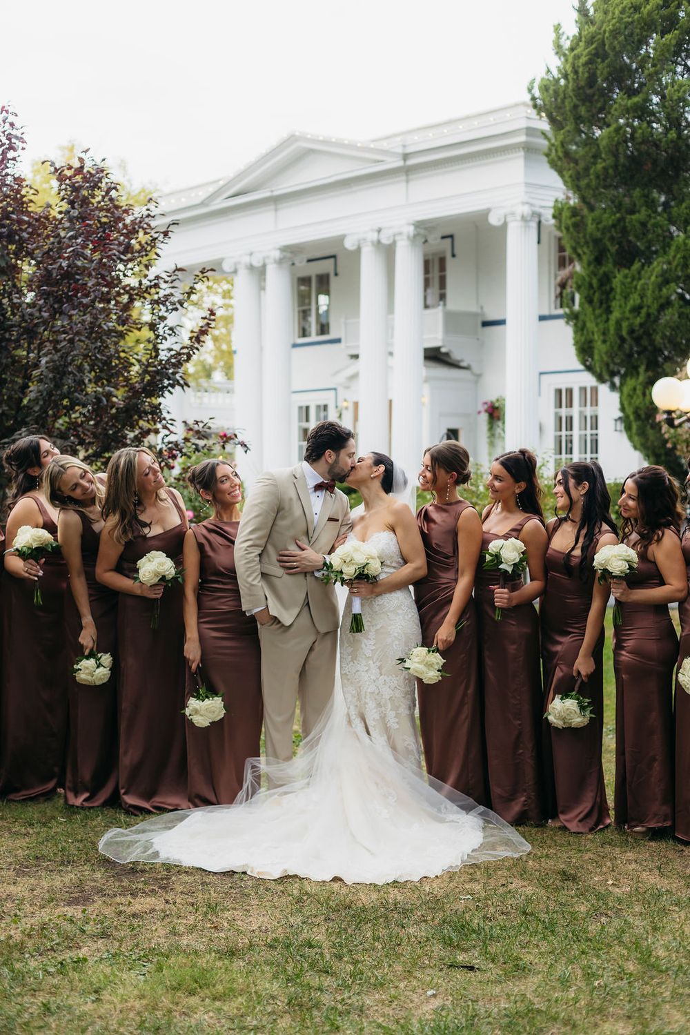 Bride and groom kissing surrounded by bridesmaids in brown gowns holding white bouquets in front of the Meson Sabika venue in Naperville, IL