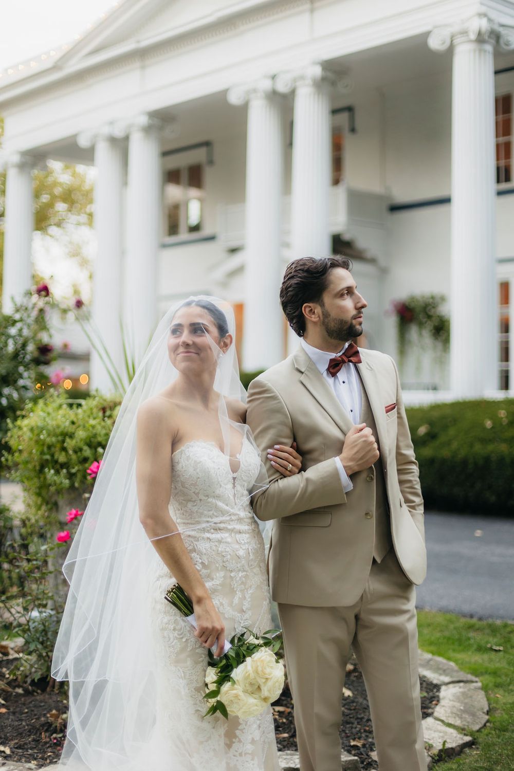 Bride in lace gown and groom in tan suit with burgundy bow tie pose in front of white columned  Meson Sabika venue in Naperville, IL