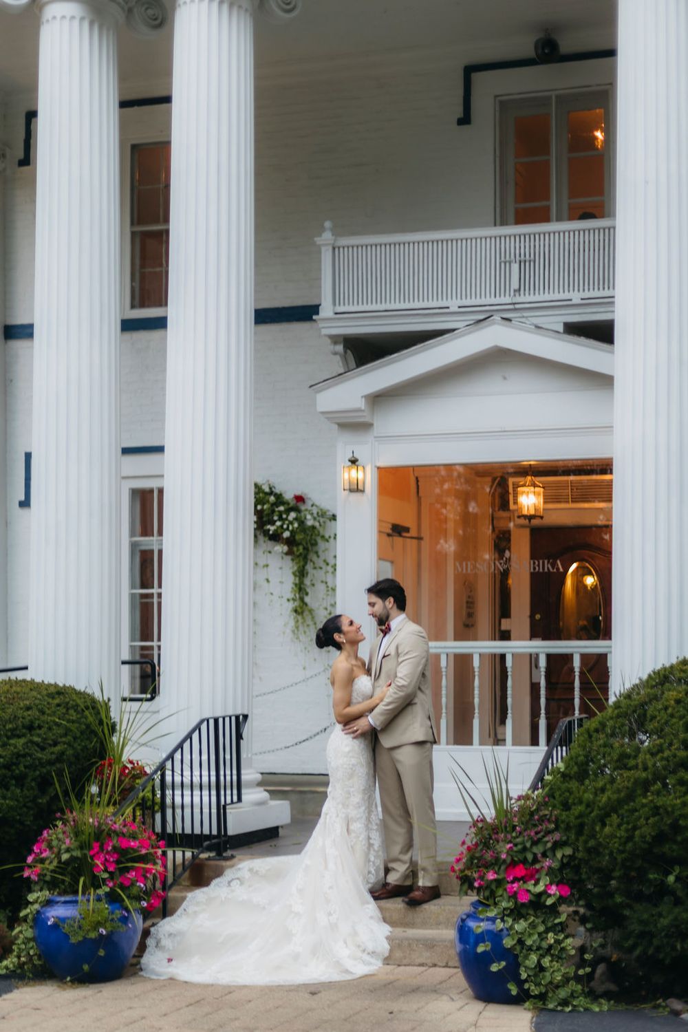 Bride and groom embrace before a grand white colonial venue Meson Sabika venue in Naperville with tall columns and colorful flowers