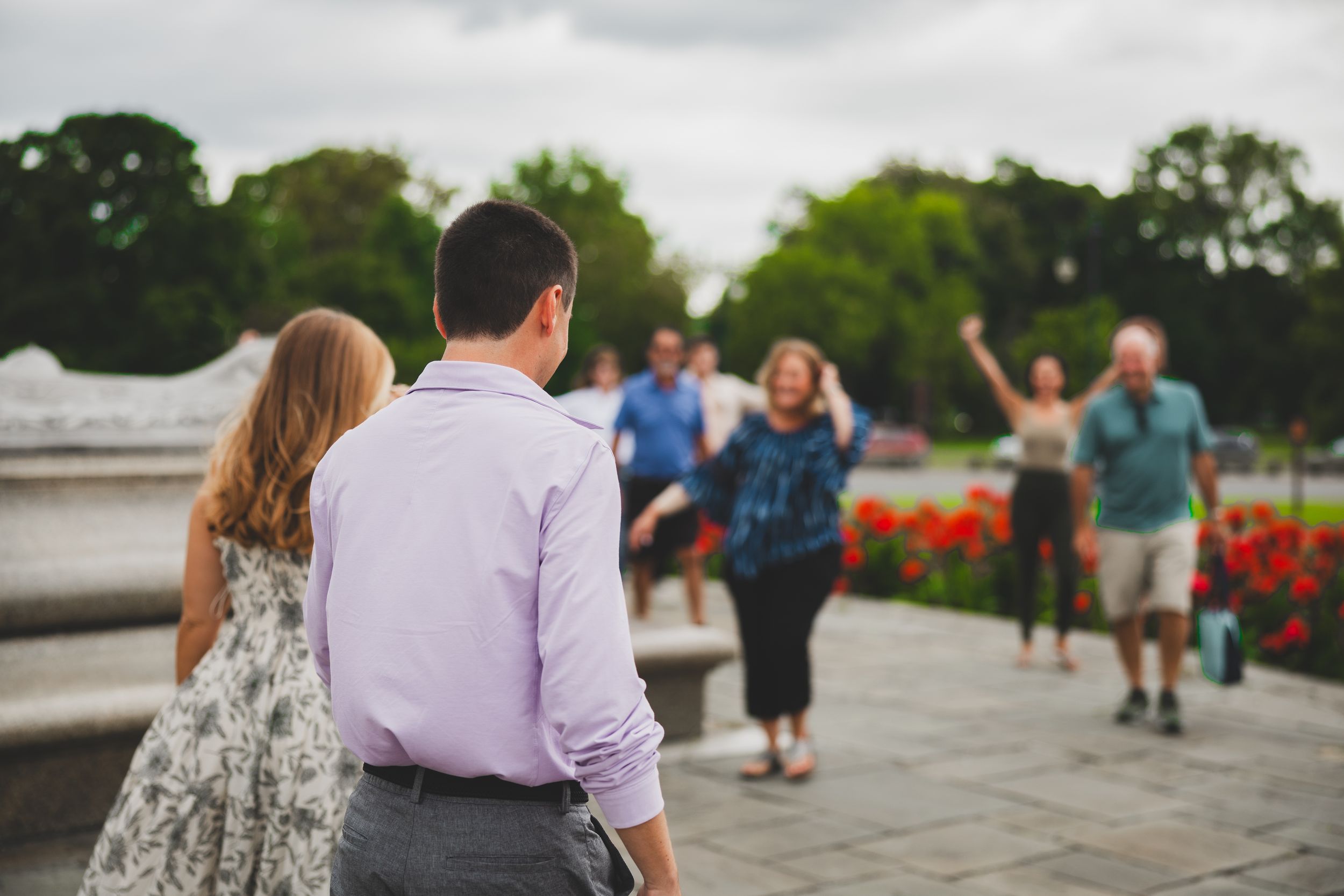 Couple watching excited friends wave in a park with red flowers and green trees in the background.