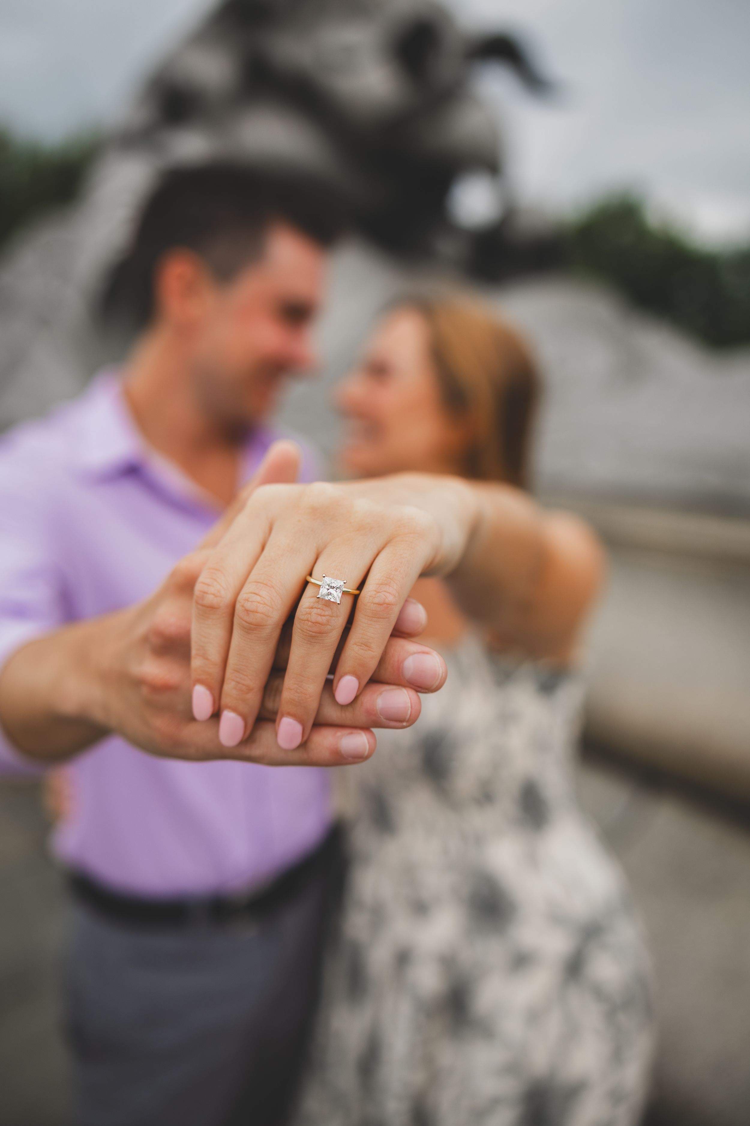 Couple holding hands close-up showing off diamond engagement ring, smiling couple blurred in background.