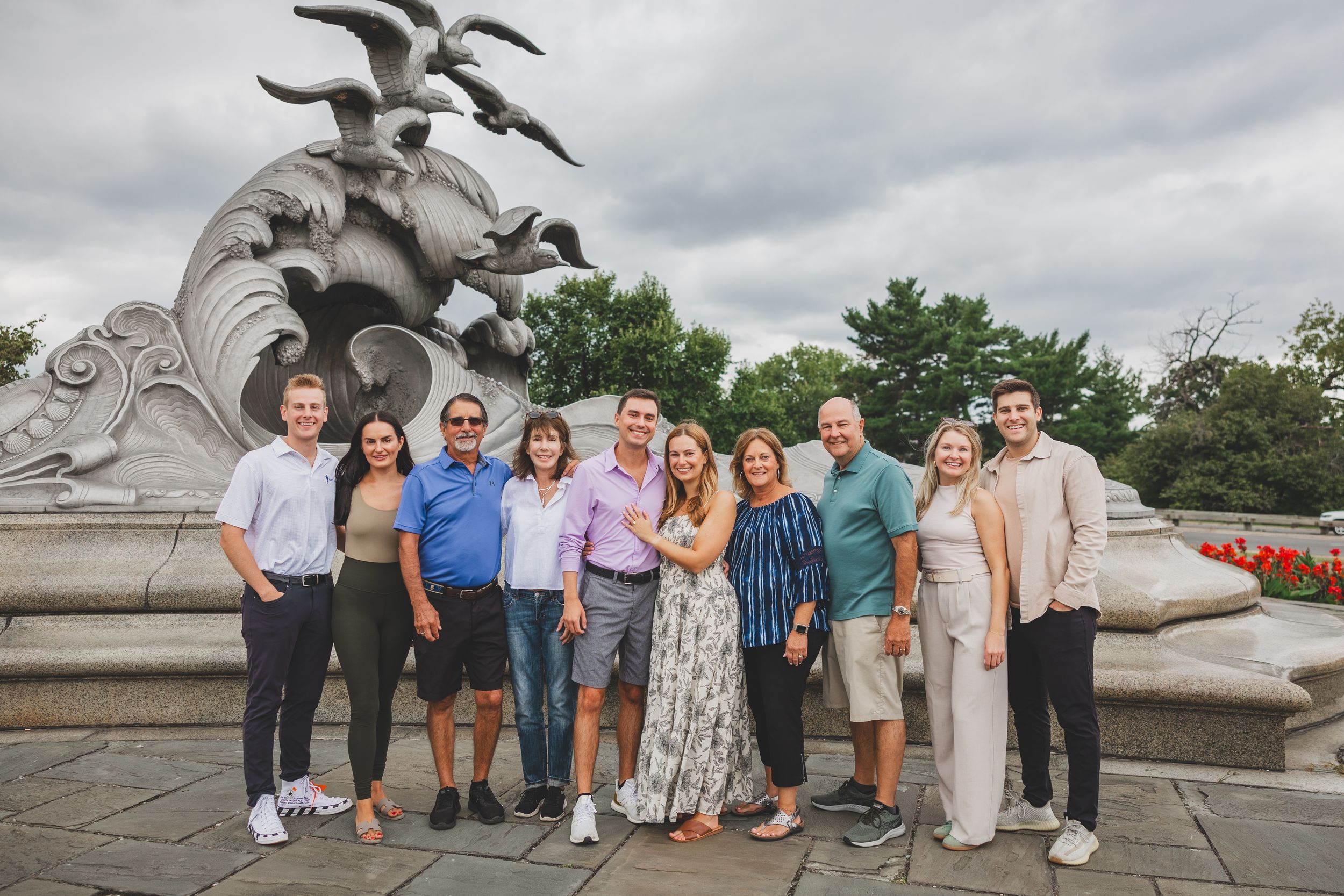 Group of ten people posing together in front of an ornate fountain sculpture on a cloudy day.