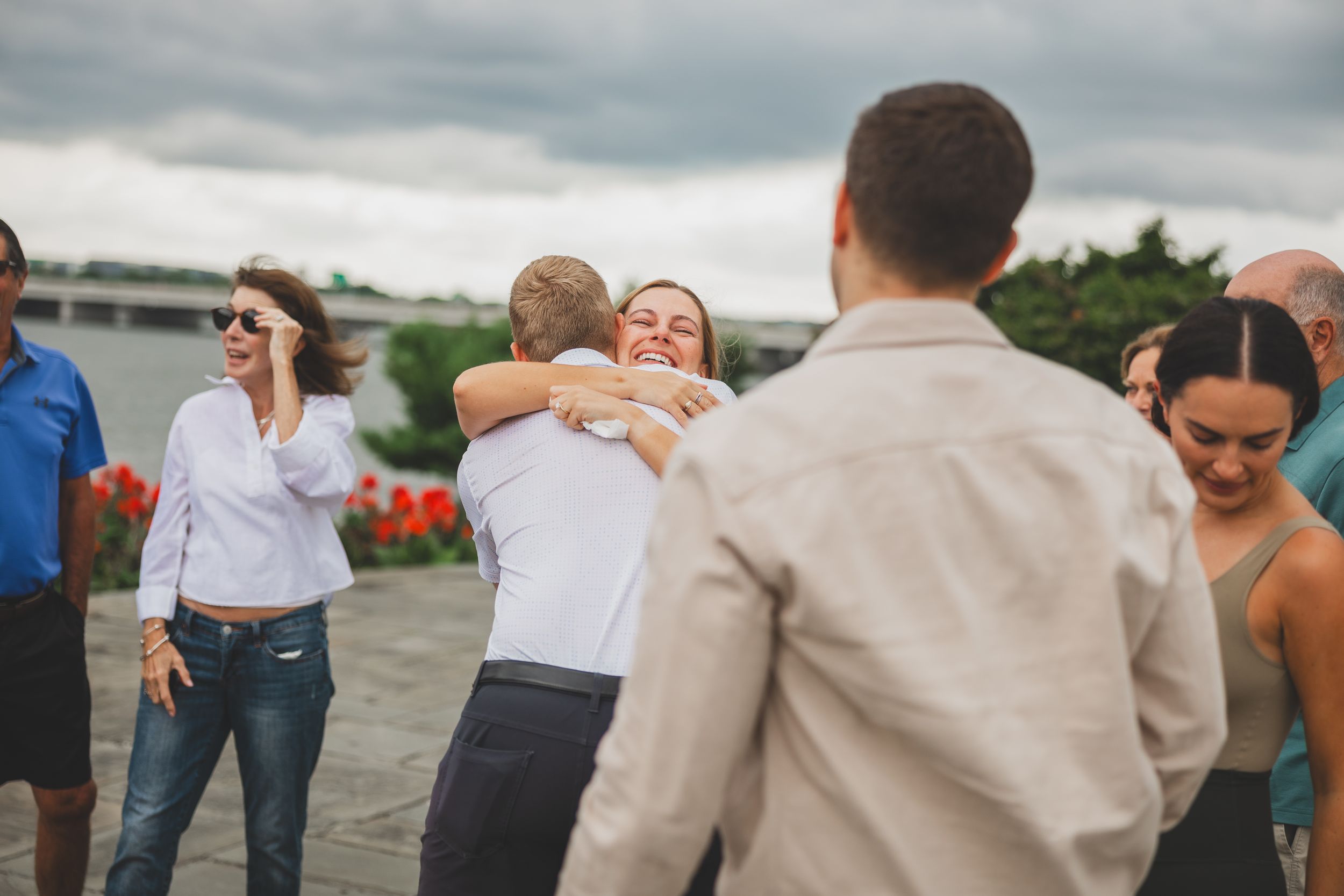 People hugging and socializing outdoors at a casual gathering under cloudy skies near a waterfront.