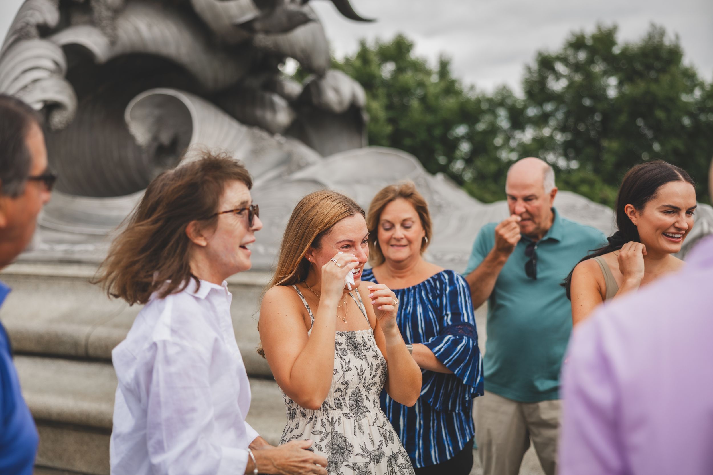 Group of people smiling and laughing outdoors near a stone monument at a casual social gathering.