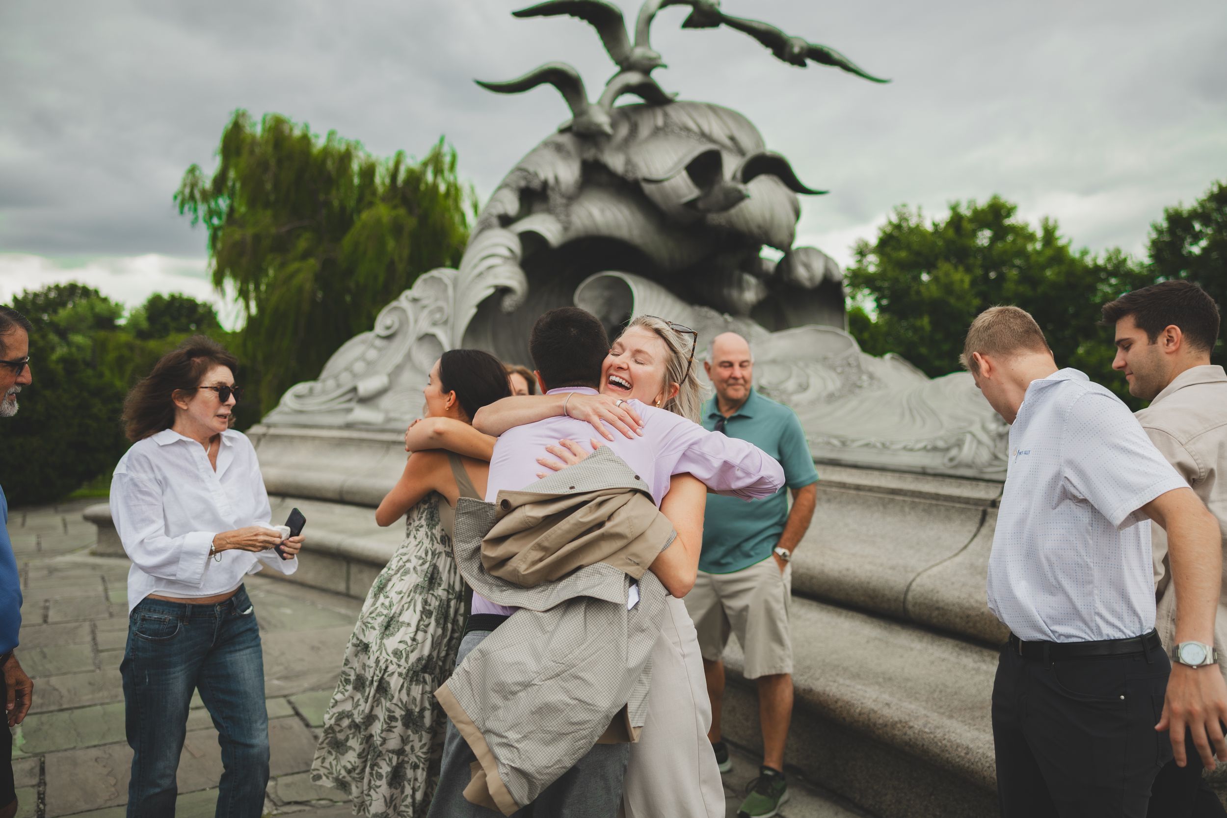 People hugging and celebrating near a stone monument sculpture in a park on a cloudy day.