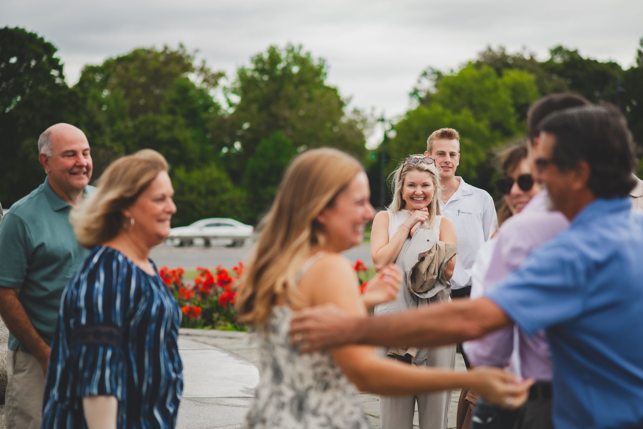 Group of people socializing outdoors at a garden party, smiling and dancing near red flowers.