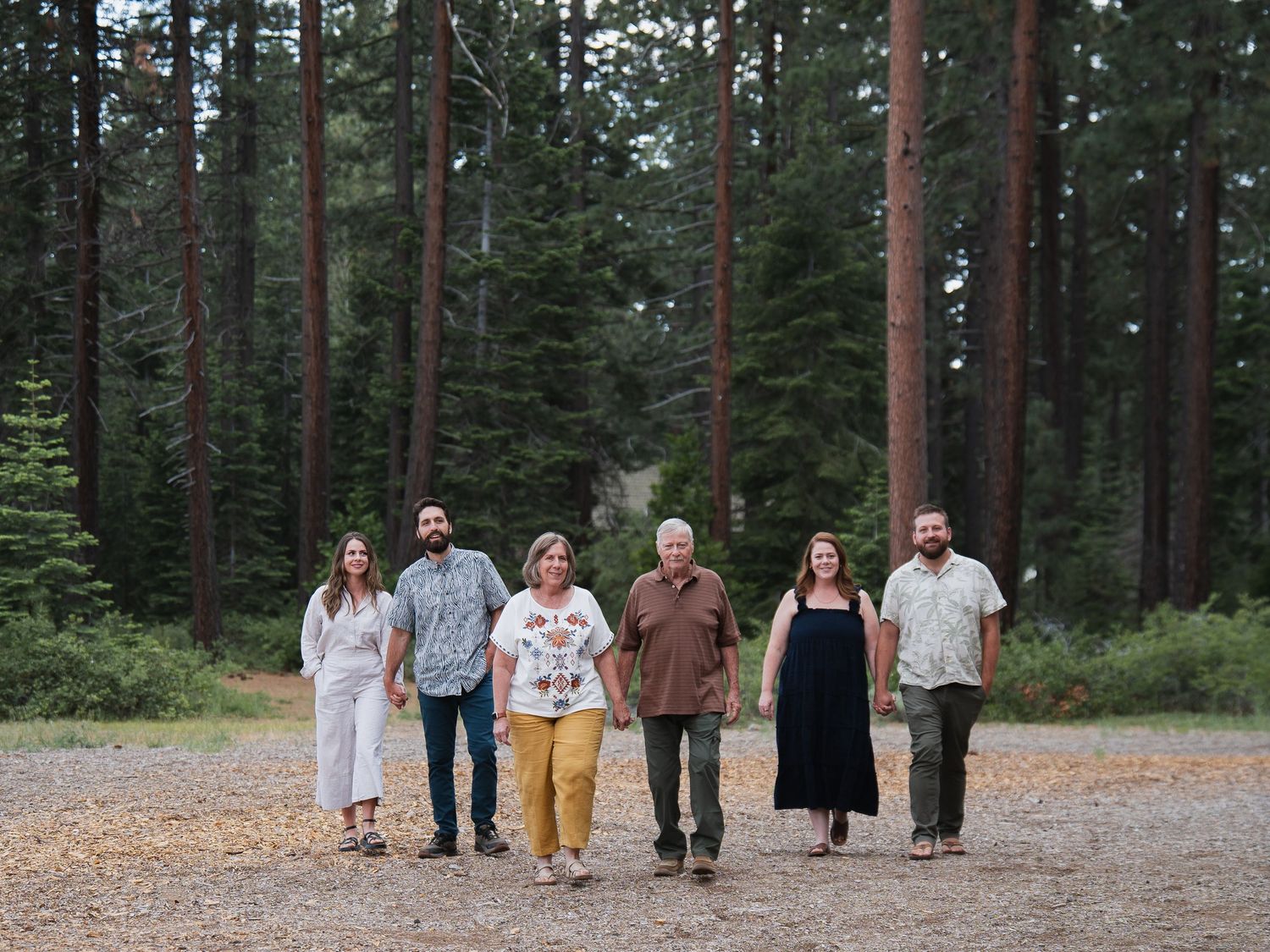 Six adults walking together on a gravel path surrounded by tall pine trees in a forest setting.