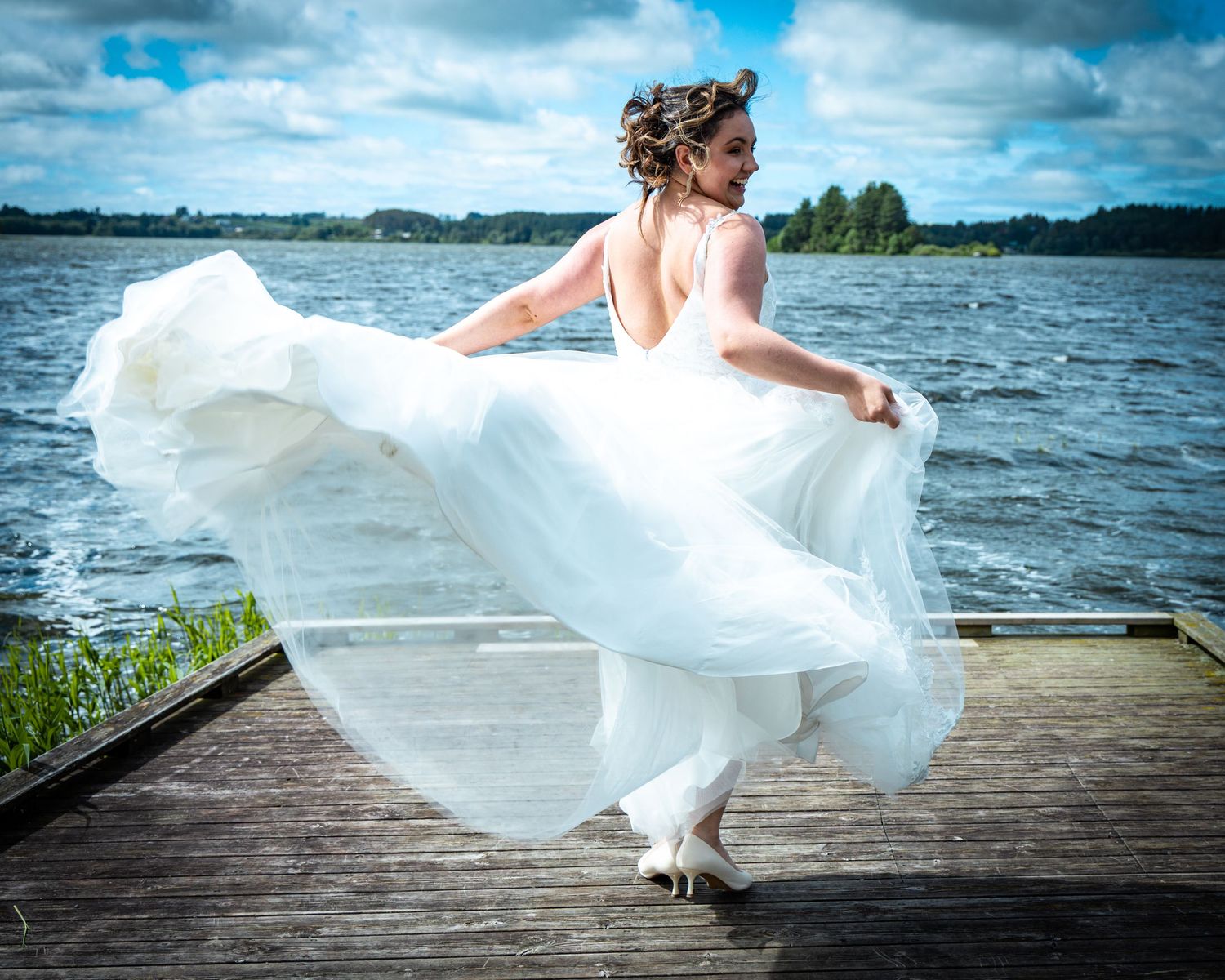 Bride in flowing white gown twirls on wooden dock beside a scenic lake under cloudy sky.