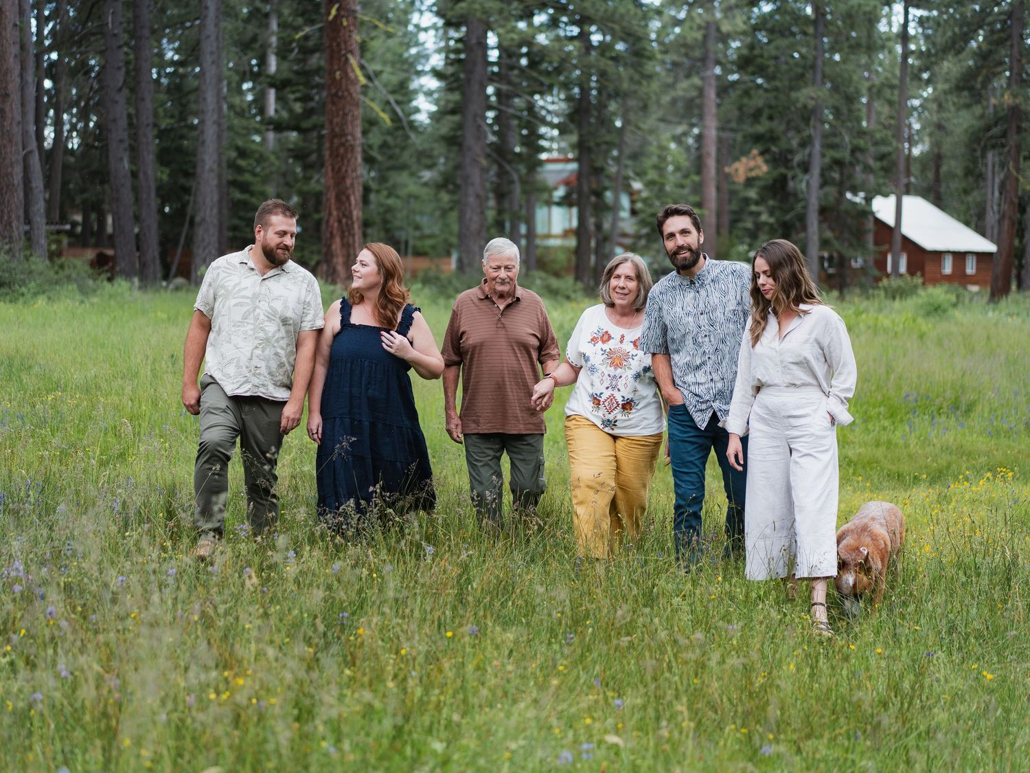 Family group of six adults walking with a golden dog in a green meadow surrounded by pine trees.