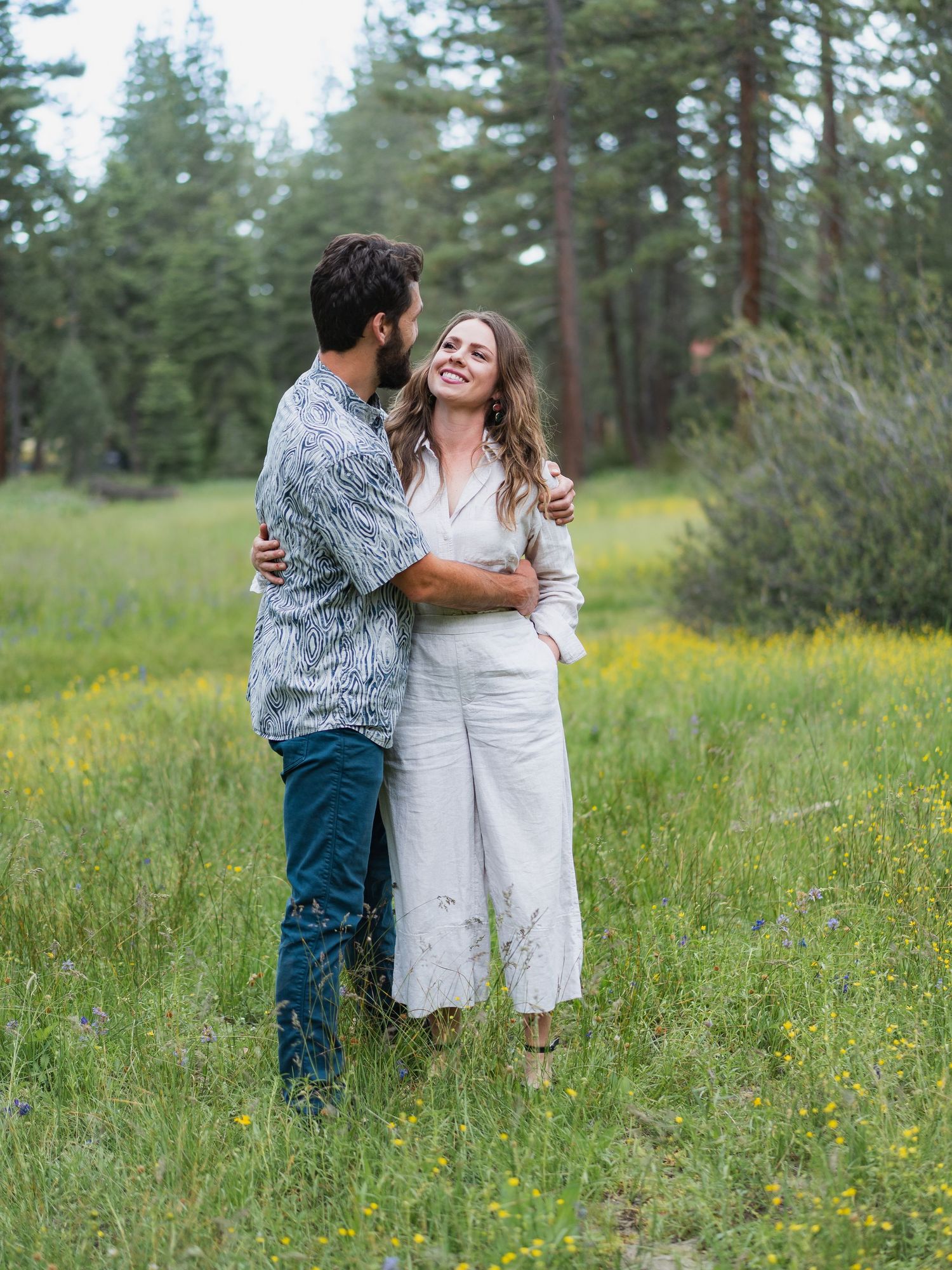Young couple embracing and smiling in a wildflower meadow surrounded by tall pine trees.