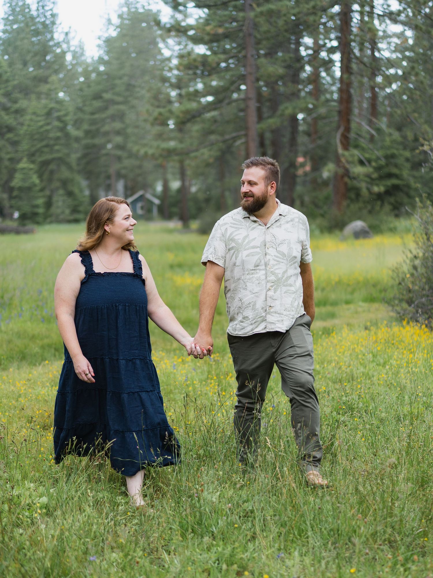 Couple holding hands in blooming meadow with pine forest backdrop.