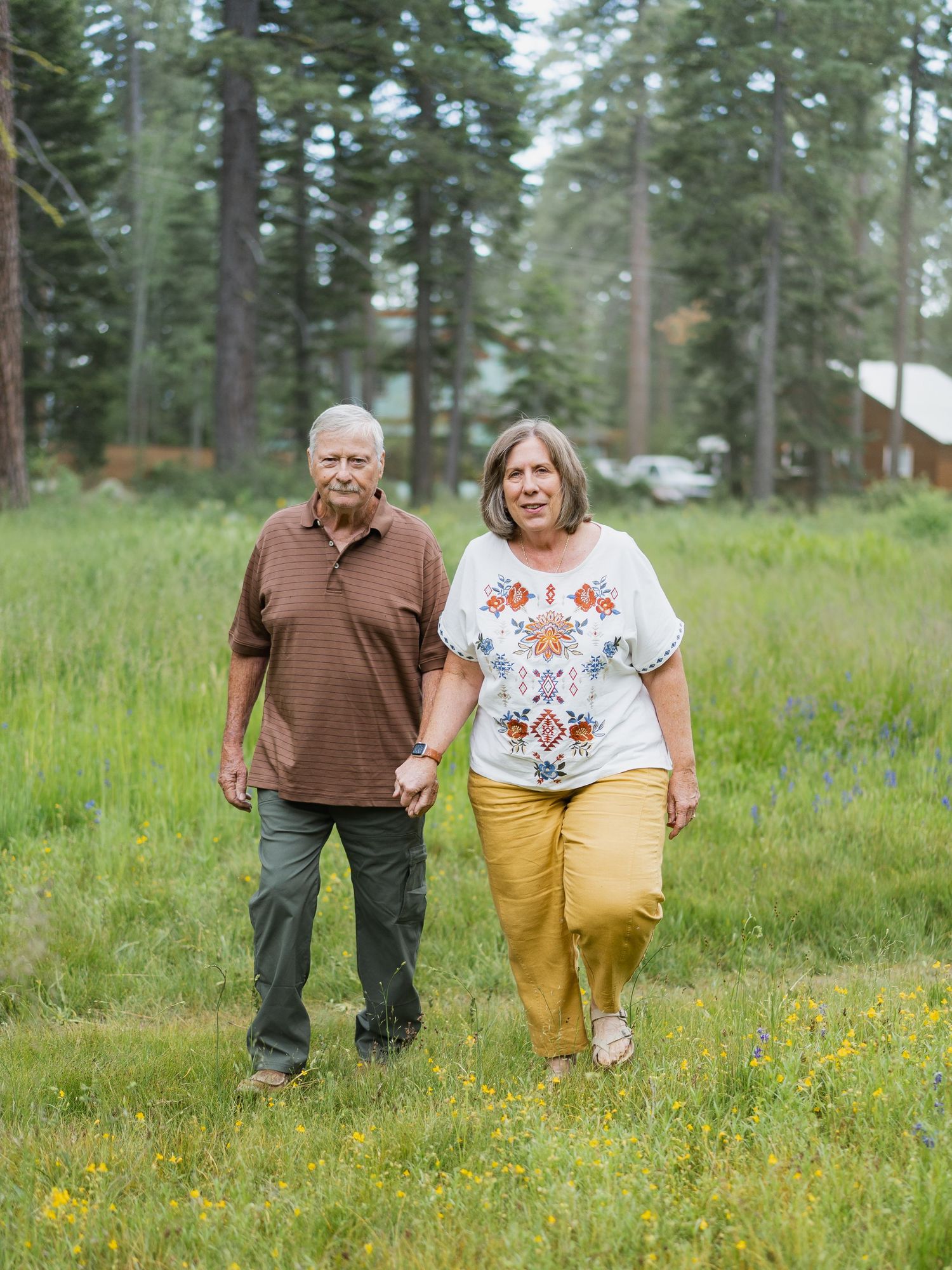 Senior couple holding hands while walking through a green meadow with pine trees behind them.