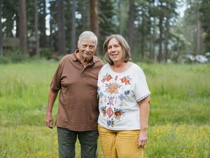 An elderly couple smiles together in a lush green meadow surrounded by tall pine trees.