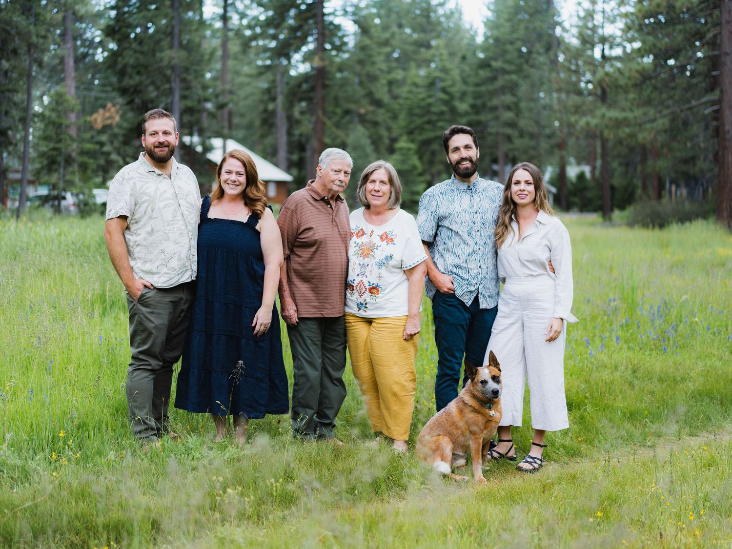 Family of six adults with a dog posing in a lush green meadow surrounded by pine trees.