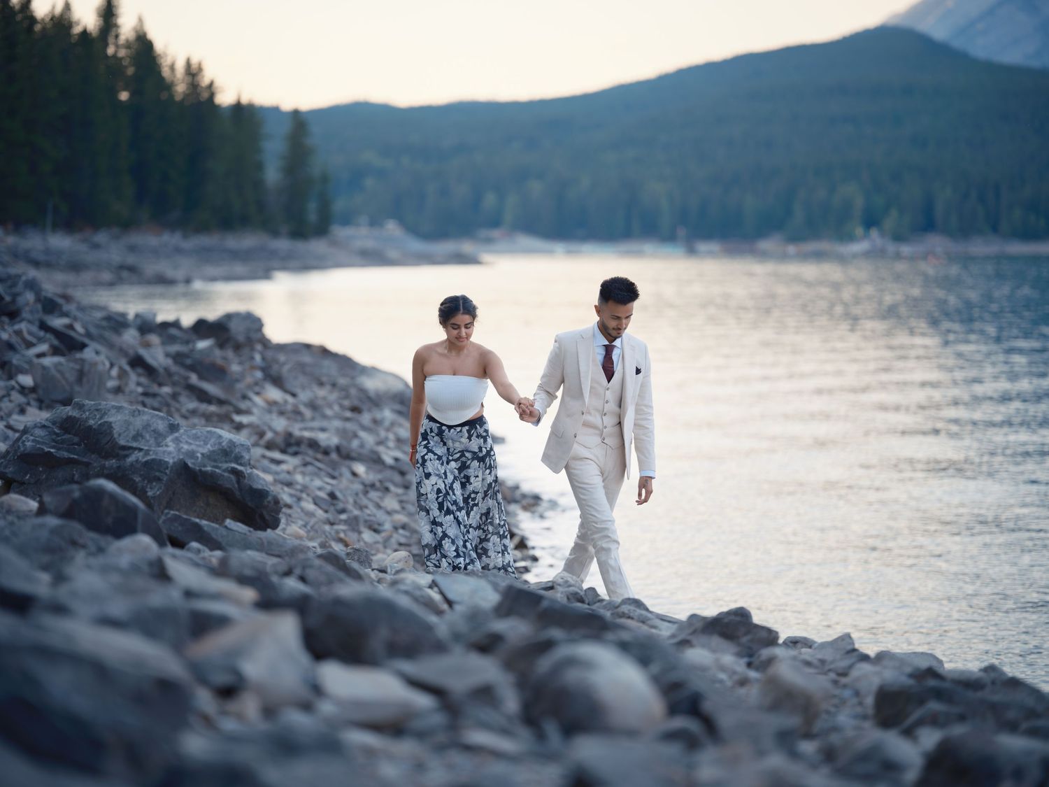 Couple walks hand-in-hand along rocky lakeshore at dusk, woman in floral dress, man in white suit, mountains behind.