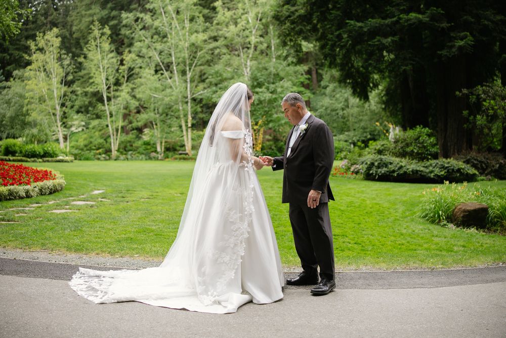 Bride in white ball gown with long veil holds hands with groom in tuxedo in lush garden setting.