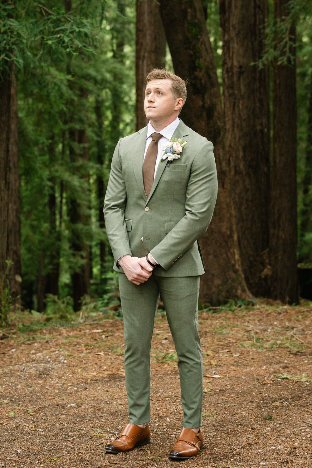Groom in sage green suit with brown tie and boutonniere standing in redwood forest.