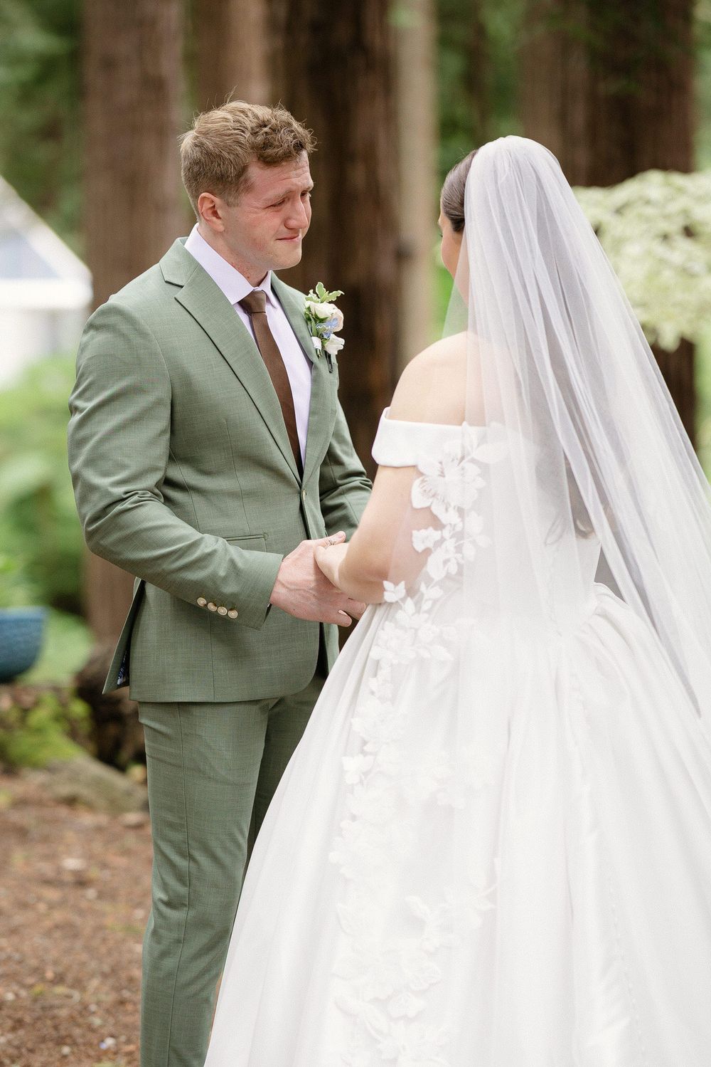 Teary-eyed groom in sage green suit holds brides hands during romantic first look in forest setting.