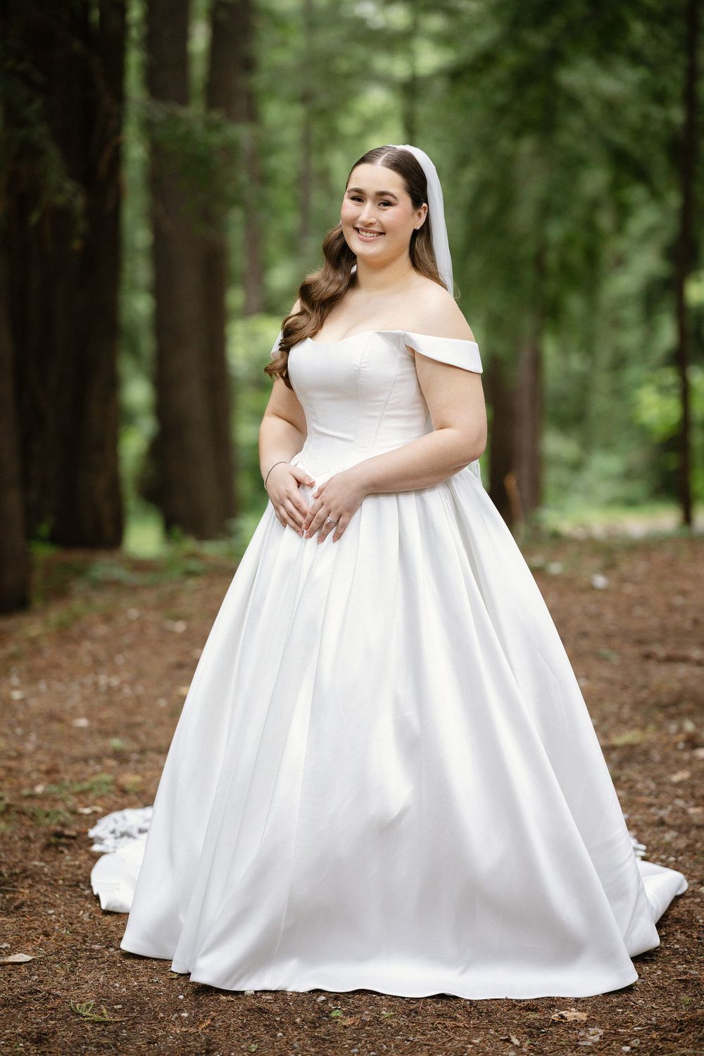 Smiling bride poses in off-shoulder white satin ball gown with veil among tall trees in forest.