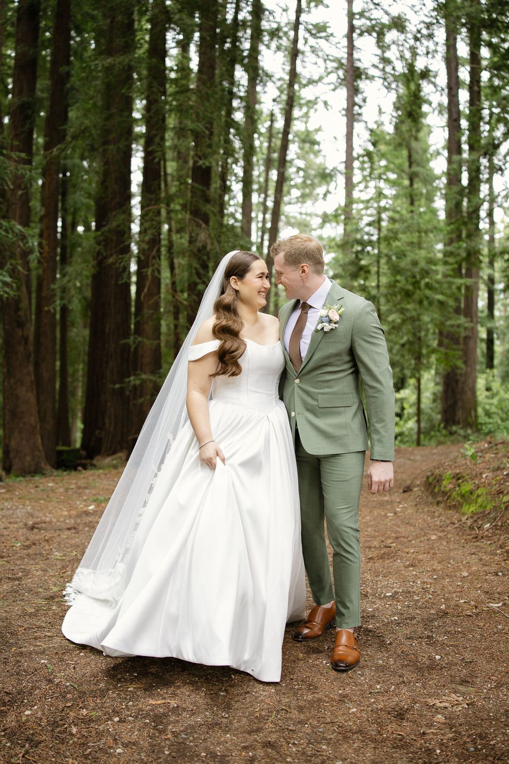 Happy bride and groom share a tender moment walking together through a lush redwood forest on wedding day.