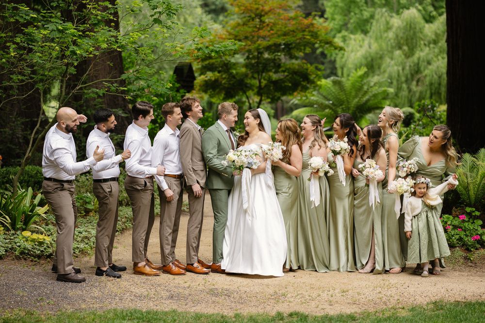 Wedding party in sage green and tan attire posing in lush garden setting.
