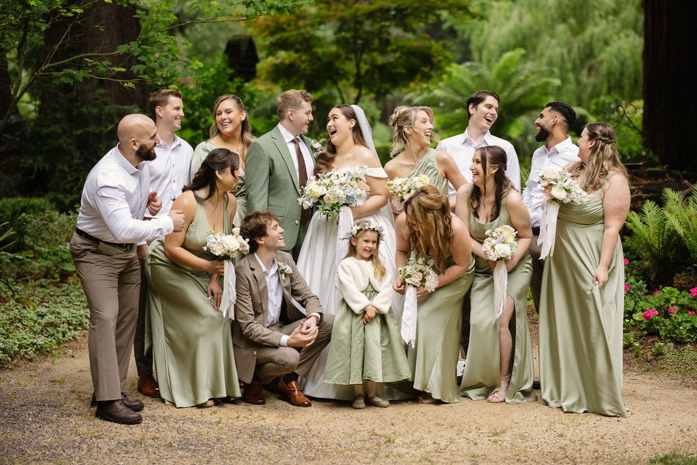 Wedding party posing in lush garden, bridesmaids in sage green gowns holding white bouquets with bride in white gown.