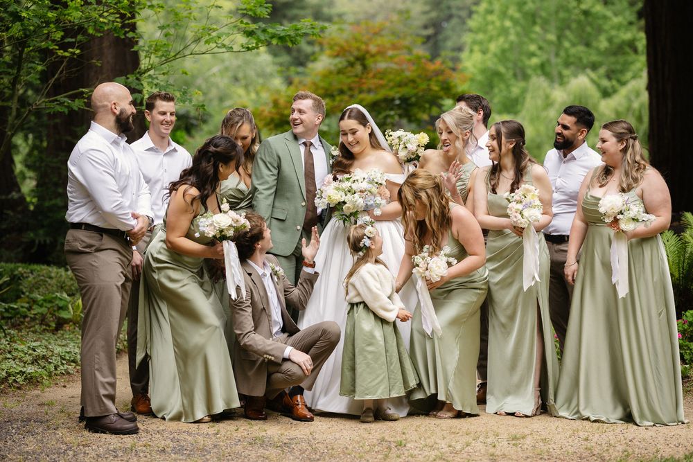 Wedding party poses outdoors in sage green bridesmaid dresses and tan suits, surrounding bride in white gown holding bouquet.