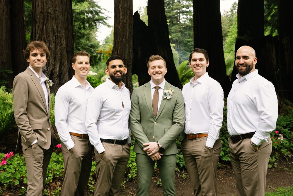 Groom in sage green suit poses with six groomsmen in white shirts and tan pants outdoors among tall redwood trees.