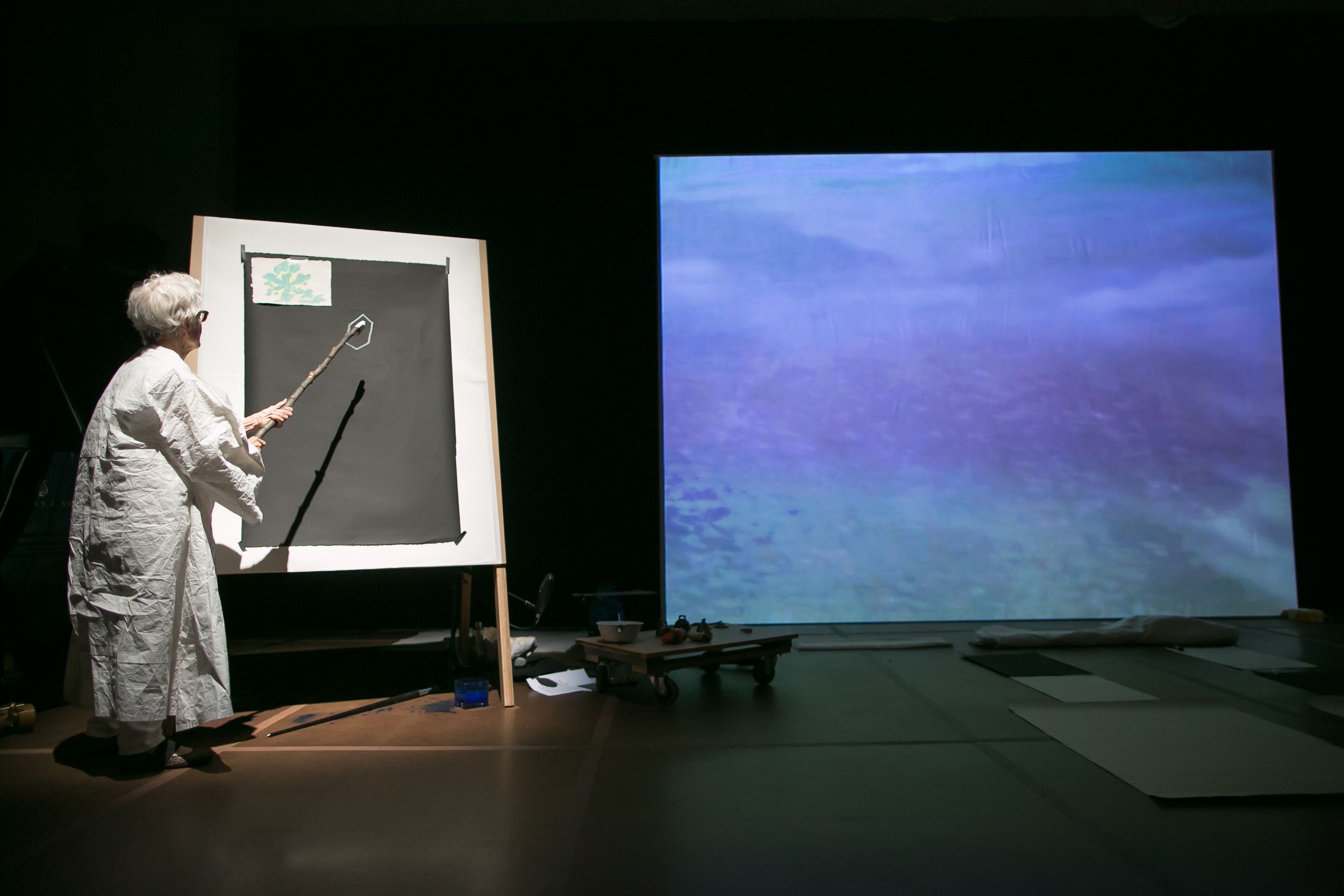 Artist in white coat pointing at canvas beside large projected ocean image on dark stage.