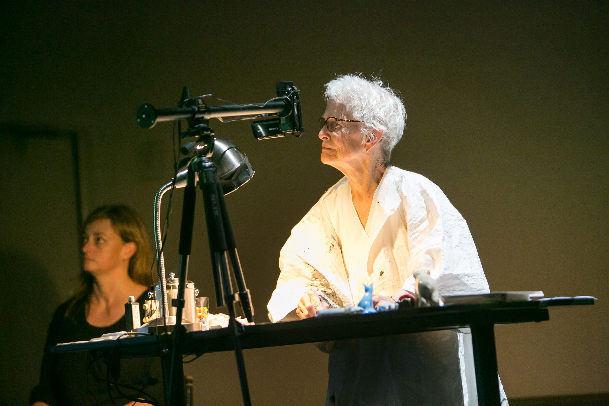 Elderly woman with white hair and glasses presenting at a desk beside a camera on a tripod during an indoor event.